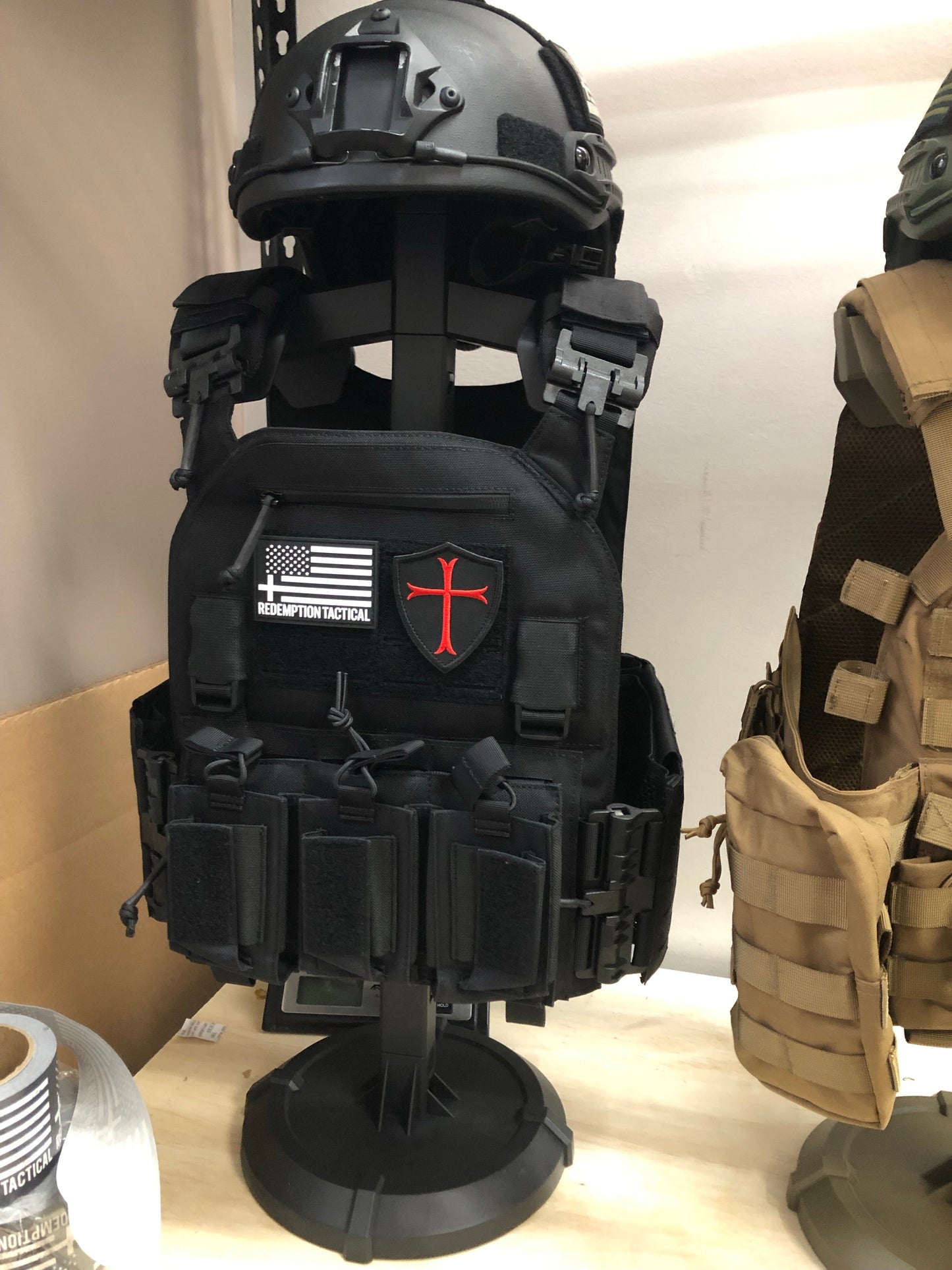 Armor Butler Stand 2.0 by Green Tip Surplus, perfect for holding full tactical vests, helmets, and battle belts. The stand is adjustable, offering flexible sizing options for various gear configurations.The image shows two tactical gear displays featuring helmets and plate carriers.
On the left is a black tactical setup with a helmet and plate carrier. The plate carrier features a patch that includes a red cross on a shield and the words "Redemption Tactical" underneath an American flag with a cross